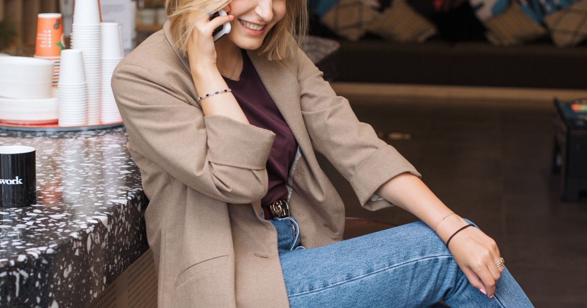 woman wears blazer with jeans; she is sitting on a stool at a coffee shop, talking on her cell phone, and laughing