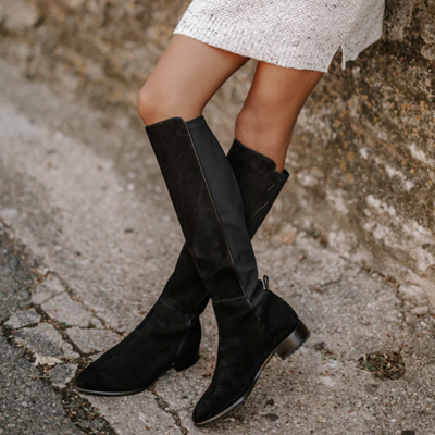 woman reclines against wall, she is wearing black knee-high boots and a beige skirt