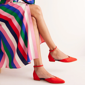 woman wears red suede ankle-strap flats with pointed toe and V on the vamp; she is wearing a stripey dress that is falling open as she crosses her legs