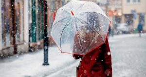 young professional woman walks along snowy city street wearing a red coat and winter business casual outfit, and carrying an umbrella