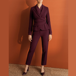 woman wears reddish suit and stands against a dark orange wall on a straw mat