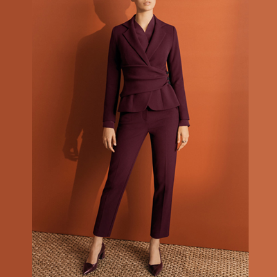 woman wears reddish suit and stands against a dark orange wall on a straw mat